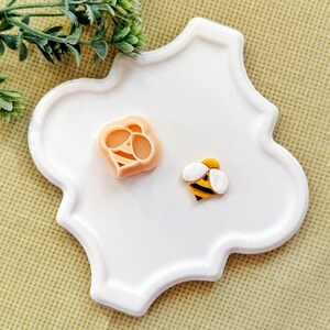 May include: A peach-colored bee-shaped cookie cutter and a small bee figurine with yellow and black stripes and white wings. The items are placed on a white, quatrefoil-shaped tray. The bee figurine features a yellow head and body.