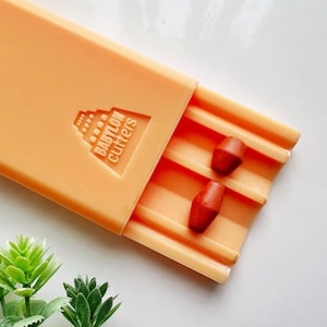 Könnte beinhalten: Orangefarbene Plastik-Pillenbox mit dem Text "BABYLON CUTTERS" auf dem Deckel. Zwei orangefarbene Pillen befinden sich in der Box.
