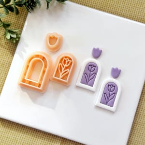 May include: A collection of polymer clay cutters and finished earrings. The cutters are peach-colored and shaped like arches and tulips. The earrings are white with purple tulip designs. The earrings have a small purple tulip at the top.