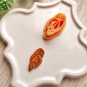 May include: Two ceramic owl figurines on a white plate. One owl is brown and the other is peach.