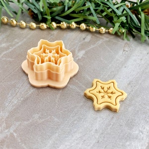 May include: A set of two beige plastic snowflake cookie cutters. The larger cutter is a multi-layered design with a snowflake pattern. The smaller cutter is a single-layer design with a snowflake pattern.