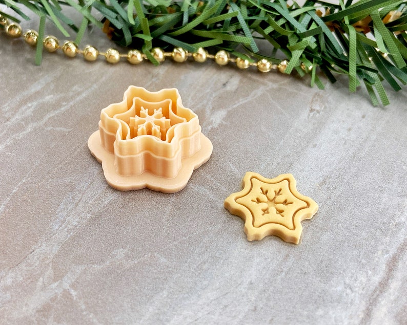 May include: A set of two beige plastic snowflake cookie cutters. The larger cutter is a multi-layered design with a snowflake pattern. The smaller cutter is a single-layer design with a snowflake pattern.