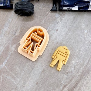 May include: Two cookie cutters in the shape of a racing suit. The larger cutter is a detailed outline of a racing suit, while the smaller cutter is a solid shape of a racing suit.
