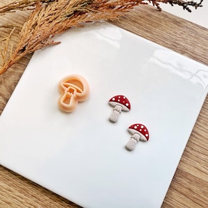 May include: A light peach-colored mushroom-shaped cookie cutter and two mushroom-shaped charms. The charms have red caps with white spots and gray stems. The items are on a white surface with a wooden background.