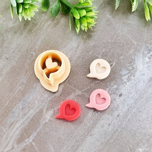 以下が含まれることがあります： ハートの入った吹き出しの形をしたクッキーカッターが4つ。カッターは薄いベージュのプラスチック製。カッターはグレーのざらざらした表面の上に置かれています。