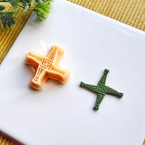 May include: Two Celtic crosses, one is a light orange colour and the other is a dark green colour. The crosses are on a white surface.