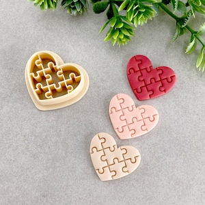 May include: A beige heart-shaped cookie cutter with a puzzle design, alongside three heart-shaped cookies in shades of pink and red. The cookies feature a puzzle piece design, perfect for Valentine's Day or other special occasions.