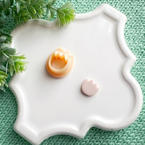 May include: Two small, decorative, resin flower shapes. One is a light orange flower with three petals and the other is a light pink flower with three petals. The flowers are on a white, scalloped, ceramic plate.