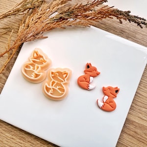 May include: Orange and white fox-shaped cookie cutters and two fox-shaped decorations on a white surface. The cookie cutters are light orange, and the decorations are orange with white bellies. A sprig of dried plants is in the background.