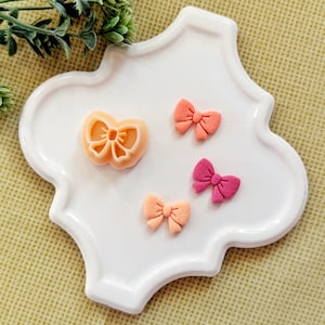 May include: A white, quatrefoil-shaped plate displays a bow-shaped cookie cutter and three small, bow-shaped decorations. The cutter and two bows are peach-coloured, while the third bow is a darker pink. The background is a light green.