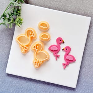 May include: A collection of flamingo-themed cookie cutters and decorations. The image shows three light orange flamingo-shaped cookie cutters, two pink flamingo decorations, and three small, round decorations. The flamingo decorations are pink with black beaks.