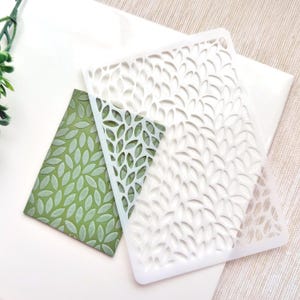 Puede incluir: Dos plantillas de plástico blanco con un patrón de hojas. La plantilla de la izquierda es más pequeña y tiene un diseño de hoja sólido. La plantilla de la derecha es más grande y tiene un diseño de hoja más intrincado.