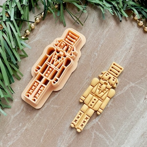 May include: Two plastic cookie cutters in the shape of a nutcracker. The larger cutter is beige and the smaller cutter is a light brown.
