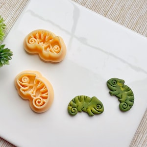 May include: Two light orange chameleon-shaped moulds and two green chameleon figurines are displayed on a white surface. The moulds have detailed carved designs. The figurines have a textured appearance and are curled.