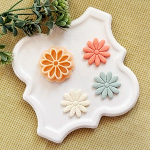 May include: A white, scalloped-edge ceramic plate holds four flower-shaped objects. One is a light orange cookie cutter, and the others are pastel-colored floral embellishments in coral, blue, and cream. Greenery is visible in the upper left corner.