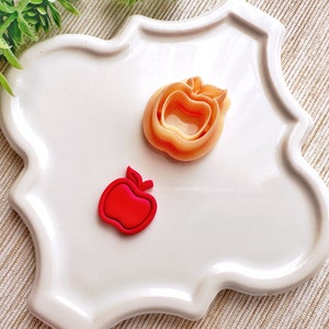 May include: Two apple-shaped cookie cutters, one red and one orange, on a white plate with a scalloped edge.