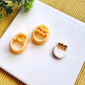 May include: Three cookie cutters in the shape of Easter eggs. Two are yellow and one is white with a brown chick inside.