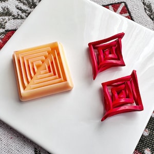 Könnte beinhalten: Zwei rote, geometrische, quadratische 3D-Designs und ein orangefarbenes, quadratisches 3D-Design mit einem Labyrinthmuster.