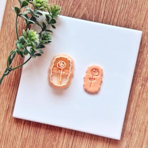 May include: An orange plastic rose-shaped cookie cutter next to a baked cookie with the same design. The cutter has a rectangular shape with a rose design in the center. The baked cookie is also rose-shaped.