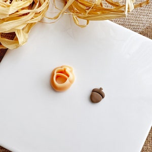 May include: Two small clay ornaments on a white surface. One is a light orange acorn with a hole in the center. The other is a brown acorn with a cap.