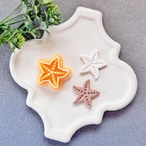 Op de afbeelding: Drie zeesterdecoraties in oranje, wit en roze liggen op een witte, decoratieve, vierlobbige plaat. De zeesterren hebben kleine stipdetails. Een takje groen gebladerte bevindt zich in de linkerbovenhoek.