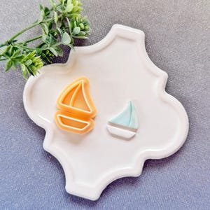 Può includere: Due formine per biscotti a forma di barca a vela, una arancione e una blu e bianca, sono su un piatto bianco decorativo. Un rametto di fogliame verde si trova nell'angolo in alto a sinistra. Le formine sono probabilmente per la cottura.