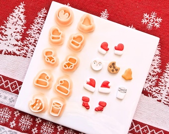 Puede incluir: Una colección de cortadores de galletas y caramelos con temática navideña dispuestos en una placa cuadrada blanca. Los cortadores tienen forma de mitones, gorros de Papá Noel y calcetines. Los caramelos incluyen un pingüino, una galleta y gorros de Papá Noel.