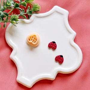 May include: A white, scalloped-edge ceramic dish holds a light orange heart-shaped object and two red ladybug-shaped embellishments with black heads and red spots. Green leafy sprigs are in the upper left corner, and the background is pink.