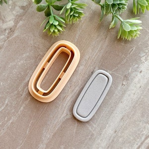 May include: Two cookie cutters, one is a double oval shape in beige and the other is a single oval shape in gray.