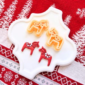 May include: A white, scalloped-edge plate holds two sets of horse-shaped items. Two are red with white floral accents, and three are tan, horse-shaped cookie cutters. The plate rests on a red and white patterned surface.