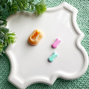 May include: Three small, colorful, bunny-shaped candies on a white plate. The candies are pink, blue, and orange.
