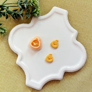 May include: A white, quatrefoil-shaped ceramic dish holds three small, yellow, cartoon-like chick figures and a peach-colored pumpkin-shaped mold. The chicks have small hats and simple facial features. The pumpkin mold has a detailed interior.