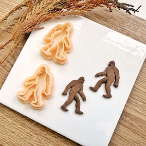 Pode incluir: Dois cortadores de biscoitos Bigfoot e dois biscoitos Bigfoot em uma superfície branca. Os cortadores de biscoitos são cor de pêssego e os biscoitos são marrons. As figuras de Bigfoot estão em poses diferentes.