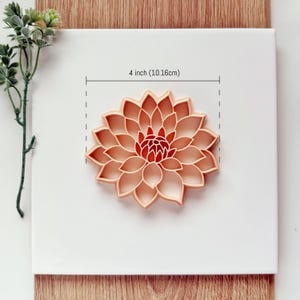 May include: A peach-colored flower-shaped cookie cutter, measuring 4 inches (10.16cm) in diameter, is centered on a white surface. The cutter has detailed petal shapes and a red center. The image includes a sprig of green leaves.