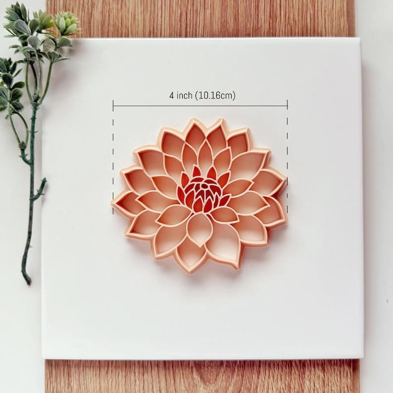 May include: A peach-colored flower-shaped cookie cutter, measuring 4 inches (10.16cm) in diameter, is centered on a white surface. The cutter has detailed petal shapes and a red center. The image includes a sprig of green leaves.