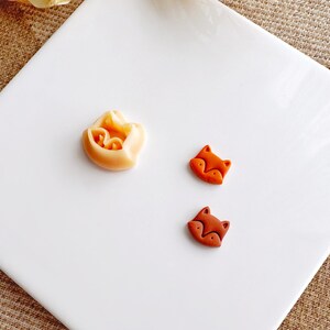 Peut inclure: Trois petites figurines de renard en argile sur une surface blanche. Un renard est de couleur orange clair et les deux autres sont de couleur orange foncé.