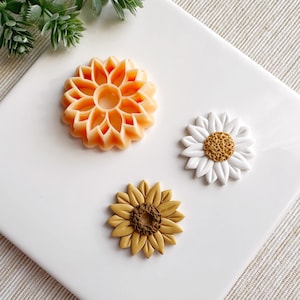 Puede incluir: Tres adornos de flores de resina en diferentes estilos. Una flor de color melocotón con un diseño geométrico, una margarita blanca con un centro amarillo y un girasol marrón con un centro amarillo.
