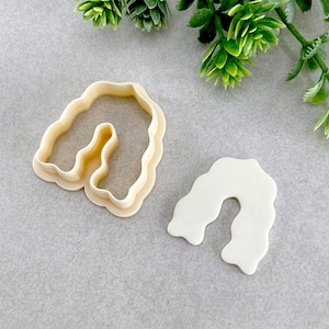 Op de afbeelding: Twee plastic koekjesvormen in de vorm van een boog. De vormen zijn beige en wit.