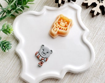 May include: Two bear-shaped items on a white, scalloped plate. One is a light orange bear-shaped cookie cutter with a scarf design. The other is a gray bear with a red and white striped scarf. The plate is on a light-colored surface.