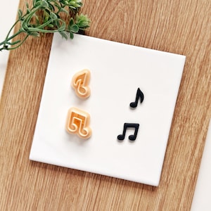 May include: Two light peach-colored plastic music note cookie cutters and two corresponding black music note shapes. The items are arranged on a white square surface. The cutters feature a single eighth note and a beamed eighth note design.
