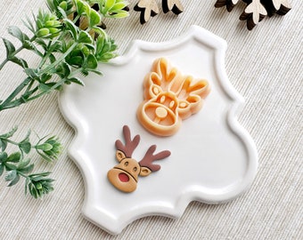 May include: Two orange plastic reindeer cookie cutters on a white ceramic plate with a scalloped edge. The reindeer have brown antlers and a red nose.