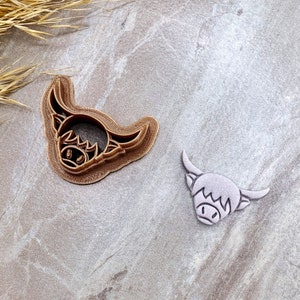 May include: Two cookie cutters in the shape of a Highland cow head. The larger cutter is brown and the smaller cutter is gray.