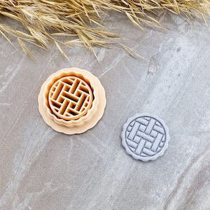 May include: Two cookie cutters, one beige and one grey, with a waffle pattern. The cutters are on a light grey surface.