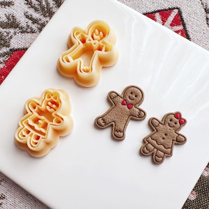 May include: Four gingerbread cookie cutters, two are brown and two are beige. The brown cutters are shaped like gingerbread people with red bows. The beige cutters are shaped like gingerbread people with a more detailed design.