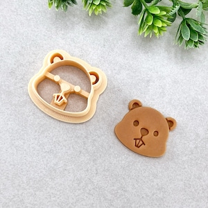 May include: A light brown cookie cutter in the shape of a cartoon beaver head, next to a baked cookie in the same shape. The cutter has a detailed outline, and the cookie has facial features.