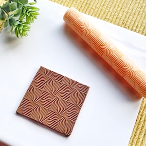 Pode incluir: Um close-up de um rolo de massa marrom e laranja com um padrão geométrico. O rolo está sobre uma superfície branca ao lado de uma peça quadrada menor de material marrom com o mesmo padrão.