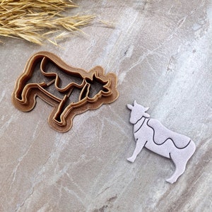 May include: Two brown plastic cookie cutters in the shape of a cow. One cutter is laying flat on a light brown surface, the other is standing upright.