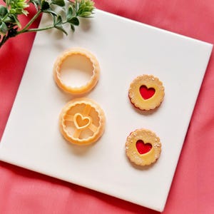 Puede incluir: Primer plano de cortadores de galletas y galletas en forma de corazón. Dos cortadores de galletas beige con bordes festoneados están junto a dos galletas en forma de corazón con relleno rojo. Las galletas están sobre una superficie blanca con fondo rosa.