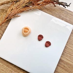 Op de afbeelding: Een lichtbeige polymeerklei eikelvormige uitsteker en twee eikelvormige kleistukken. De uitsteker heeft een afgeronde vorm met een centrale opening. De eikels zijn bruin en roodbruin. De items liggen op een wit oppervlak.