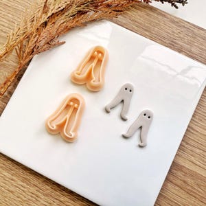 Pode incluir: Uma coleção de cortadores de biscoitos e decorações de fondant com tema de Halloween. Os cortadores têm o formato de fantasmas, dois em cor pêssego claro e dois em cinza. As decorações são pequenas, com olhos simples de pontos pretos.